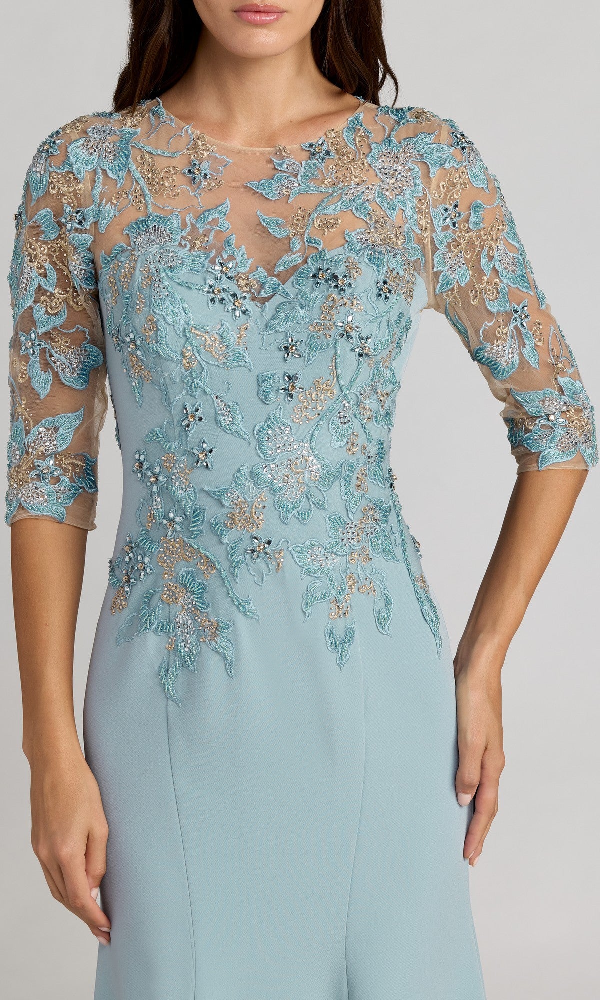A woman is wearing the Mac Duggal 13204 long formal dress in light blue, featuring sheer floral lace and bead embellishments on the bodice and sleeves, while standing against a plain background.